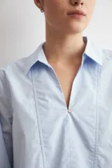 Blusa de algodón color celeste, con cuello camisero, escote en V y mangas largas con puños anchos. Presenta un diseño de corte recto y holgado.