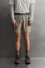 Pantalón corto de trail running color beige, confeccionado en tejidos técnicos ligeros y elásticos. Cuenta con forro interior, cintura elástica ajustable con cordón, bolsillos traseros laterales multifunción, bolsillo trasero central con cremallera, bajo con costura termofijada y aberturas laterales. Incluye logo estampado en la pernera.
