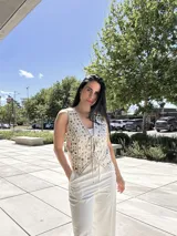 Chaleco sin mangas color beige claro, con escote en V y cierre frontal mediante cordones anudados. Presenta un diseño troquelado con apliques circulares y romboidales en tonos beige y plateado/azul claro.