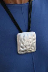 Collar de diseño moderno con dije cuadrado de superficie irregular y acabado plateado mate, suspendido de múltiples cordones negros finos.