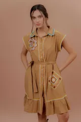 Vestido midi de color tostado confeccionado en algodón, con diseño camisero de manga corta, cuello camisero, botonadura frontal y cinturón ajustable en la cintura. Presenta detalles de bordados geométricos y florales en contraste, además de apliques de encaje y un volado en el ruedo.