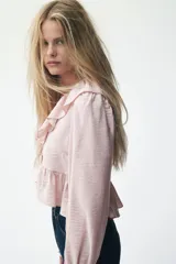 Blusa corta color rosa pálido con tejido suave y leve brillo. Presenta un escote en V pronunciado adornado con un ribete ancho de volados que continúa en el bajo de la prenda. Tiene mangas largas con volumen y puños con volado.