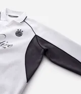 Buzo deportivo de felpa en color blanco con detalles en negro. Presenta cuello tipo polo, mangas largas con puños acanalados y diseño con recortes laterales. Incluye estampados gráficos en el frente con tipografía cursiva y emblema circular.