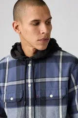 Camisa de franela a cuadros en tonos azul, gris y blanco, con capucha negra y cordones ajustables. Presenta dos bolsillos frontales con solapa y botones, y cierre de botones en el frente.