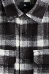 Sobrecamisa de franela con diseño a cuadros en tonos marrón oscuro y blanco hueso. Presenta cuello inglés, tapeta frontal con botones, dos bolsillos superiores con solapa y mangas largas con puños abotonados.