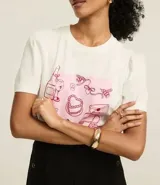 Remera blanca de algodón con cuello redondo y mangas cortas abullonadas. Presenta un estampado con dibujos de objetos femeninos en tonos rosados.