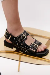 Sandalias negras de cuero con suela plana, dos tiras anchas con hebillas doradas y apliques metálicos.