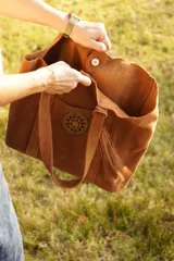 Cartera tipo tote de gamuza color marrón, con diseño amplio y relajado. Presenta un bolsillo frontal externo con cierre de broche metálico y una hebilla decorativa circular, además de un detalle de borla colgante en el lateral.