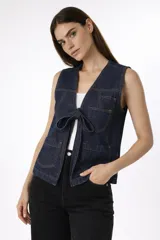 Chaleco de denim azul oscuro con cierre frontal mediante lazo, bolsillos delanteros tipo parche y costuras a la vista.