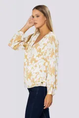 Blusa de manga larga con estampado floral en tonos blanco y mostaza. Presenta escote en V con botones frontales, frunces en los hombros y puños abotonados con detalle de pin metálico con logo.