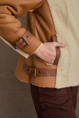 Chaqueta tipo gamulán de cuero color camel con interior y solapas de piel sintética en color beige. Presenta cierre frontal cruzado, correas ajustables con hebillas en los puños y en la cintura, y diseño de cuello solapa.