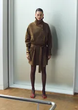 Chaqueta de gabardina color verde militar, de corte oversize con cuello alto abotonado de manera asimétrica, mangas largas con puños ajustables y cinturón ajustable del mismo tejido.