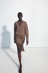 Vestido corto marrón con rayas verticales blancas, cuello camisero, manga larga y detalle de nudo y lazada en la falda. Cuenta con cierre frontal de botones.