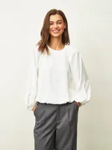 Blusa de color blanco hueso con cuello redondo, mangas largas abullonadas y puños elásticos. Presenta un diseño con frunces en los hombros y ruedo elástico que le otorga una caída holgada.