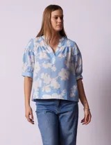 Blusa de gasa estampada con flores blancas sobre fondo celeste, marca Floral & Ivy. Cuello a la base con escote en V. Manga corta con detalle de puño y botón.