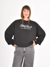 Buzo negro de corte holgado con cuello redondo y mangas largas con puños elásticos. Presenta un estampado frontal con la frase 'Shine Bright' en tipografía cursiva y texto complementario en blanco.