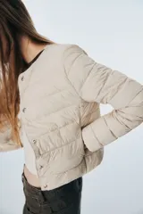 Campera acolchada de nylon color beige, con cuello redondo, cierre de botones a presión y bolsillos laterales discretos. Tiene elástico fino en los puños y está forrada.
