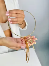 Colgante de diseño minimalista compuesto por una estructura circular rígida de metal dorado y un dije central en forma de estrella o destello de líneas rectas. Incluye tiras de cuero marrón con detalles metálicos que aportan un toque artesanal.