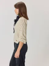 Buzo de lana color beige con mangas abullonadas de tejido calado con diseño de rombos. Presenta un cuello redondo y un detalle de flor negra de tela con cintas colgantes en el escote.