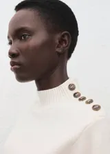 Jersey de punto medio color blanco, con cuello perkins y detalle de botones metálicos en el hombro izquierdo.