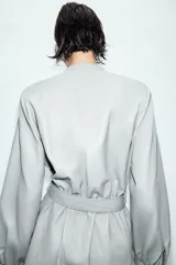 Vestido corto de efecto piel en color celeste grisáceo, con cuello redondo, manga larga con puños elásticos y hombreras. Presenta un cierre frontal con botones a presión y un cinturón ajustable del mismo tejido con hebilla metálica.
