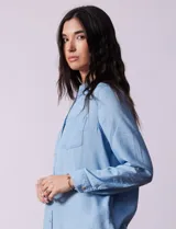 Camisa celeste de lyocell, con cuello a la base, escote en V y abertura central con botones. Tiene bolsillos aplicados en la parte delantera y mangas 3/4 regulables con presilla y botón.