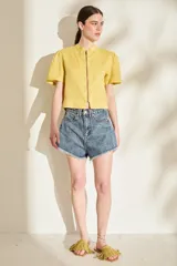 Blusa de lino color amarillo, con cuello mao, cierre frontal con cremallera y mangas cortas abullonadas. Cuenta con un bolsillo aplicado en el frente.