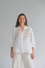 Blusa blanca de corte holgado con cuello mao y cierre frontal de botones. Presenta mangas largas abullonadas con puños elásticos y un delicado bordado en color beige en la parte superior delantera.