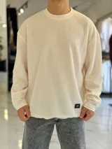 Buzo oversize de tejido waffle en color beige, con cuello redondo y manga larga, con pequeña etiqueta de marca en el dobladillo inferior.