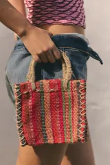Mini bolso cuadrado de yute 100%, con diseño de rayas multicolor en tonos rojos, verdes y tostados. Cuenta con asas de mano rígidas forradas en yute y costuras decorativas entrelazadas en los laterales.