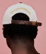 Gorra de algodón color blanco con visera curva y ajuste de hebilla de cuero marrón. Presenta bordado frontal con letras "Pagodinho F.C." en dorado.
