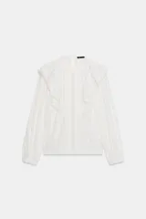 Camisa blanca de cuello redondo y manga larga, con detalle de volantes frontales y entredoses de encaje. Presenta un cierre en la espalda con abertura y botón.