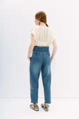 Pantalón de denim azul de tiro alto con cintura estilo paper bag y lazo a tono. Presenta un bordado elaborado con lentejuelas y piedras en forma de palmeras en la parte lateral de la pierna.