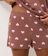 Pijama corto de dos piezas compuesto por una remera de manga corta con cuello redondo y bolsillo frontal, y un short de cintura elastizada. Ambos presentan un diseño estampado de corazones sobre fondo marrón.