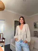 Camisa de color beige con escote en V, mangas largas abullonadas y detalle de frunces con cordones ajustables en la parte frontal.