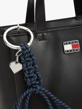 Cartera tote de poliuretano sintético liso color negro de la marca Tommy Jeans, con dos asas superiores y correa ajustable. Presenta cierre de cremallera, compartimento principal e insignia metálica de la marca en la parte delantera. Incluye un adorno colgante extraíble con cordón azul trenzado, dije en forma de corazón y herrajes plateados.