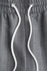 Pantalón de vestir gris oscuro con finas rayas verticales blancas (pinstripe), tiro alto y cintura elástica con cordón blanco ajustable. Presenta un corte ancho y recto, con una fina franja blanca lateral que recorre la pierna.