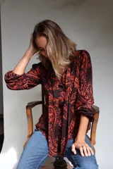 Camisa de lino con estampado paisley en tonos marrón y naranja, cuello con lazos integrados, mangas largas con frunces en hombros y puños elásticos, y cierre frontal con botones de coco.