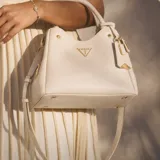 Cartera tipo bolso de mano Guess modelo Meridian II, color blanco, con textura granulada. Presenta herrajes dorados, logo triangular metálico en el frente y colgante decorativo. Incluye asas superiores fijas y correa larga ajustable extraíble.