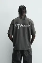 Camiseta de manga corta y cuello redondo, color gris oscuro o negro deslavado, con estampado de texto en blanco en el pecho.