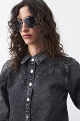 Camisa de jean negra con efecto lavado, cuello clásico, cierre frontal con botones plateados y detalles de tachas metálicas en el canesú y el frente.