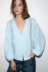 Blusa de escote en pico con cierre frontal de botones, mangas largas abullonadas con bordados a contraste y detalles de encaje tipo entredós y jaretas en el frente.