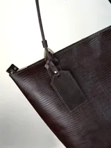 Cartera tipo tote de cuero genuino color marrón oscuro con textura de reptil. Cuenta con doble asa de hombro, cierre superior y un dije colgante de cuero a tono.