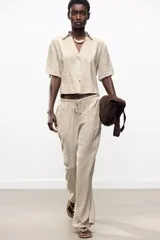 Pantalón negro de tiro alto con cintura elástica ajustable mediante cordones con terminales dorados. Presenta pernera ancha y bolsillos laterales. La tela tiene una textura sutilmente arrugada o plisada.