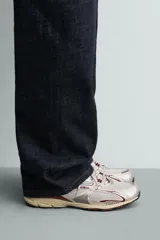 Zapatilla deportiva tipo running con diseño técnico, confeccionada en una combinación de materiales sintéticos y malla. Presenta una estética metalizada en tonos plata con detalles en color granate y una suela con volumen de diseño irregular en tono crema.