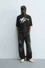 Camiseta negra de corte relaxed fit, cuello redondo y manga corta. Presenta un estampado frontal en blanco con efecto spray que simula dos figuras volando, y texto en un óvalo que dice "SUCCESS CHASER".