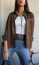 Camisa de gamuza color chocolate con bordados decorativos en color crudo a lo largo de la botonadura frontal y en el bolsillo superior. Presenta cuello clásico, cierre frontal con botones y mangas largas.