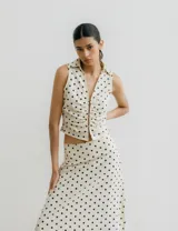 Chaleco blanco con estampado de lunares negros, cuello camisero y cierre frontal con broches metálicos.