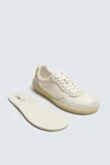 Zapatilla deportiva tipo barefoot confeccionada en piel color blanco roto con apliques de gamuza en tonos grises. Posee puntera redondeada y ancha, cierre mediante cordones al tono y suela de goma flexible en color caramelo con tecnología drop 0.