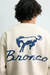 Sudadera de tejido con algodón, color beige claro, de corte relajado (relaxed fit) y cuello redondo. Presenta un parche bordado en el pecho con el logo de Ford y el texto "Genuine Parts" en azul marino.